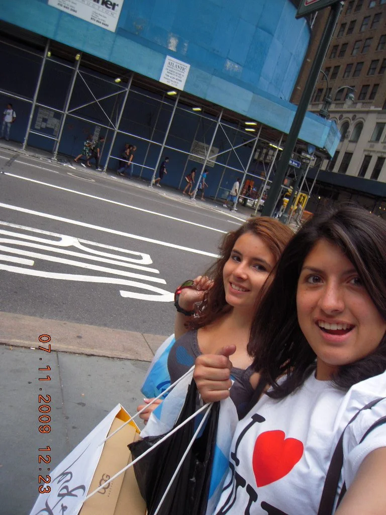 Two young women taking a selfie on a city sidewalk with a construction site and pedestrians in the background.