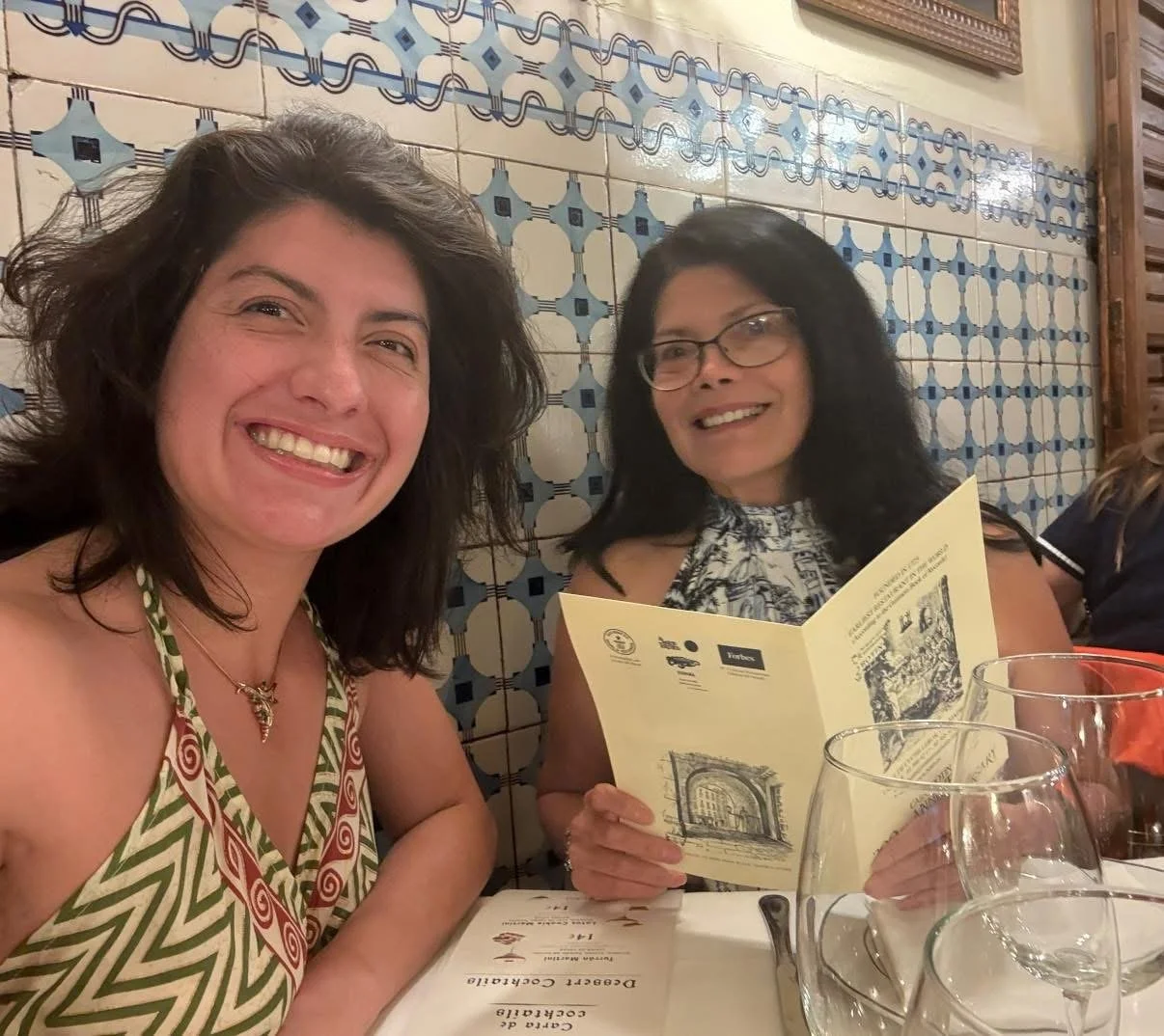 Two women sitting at a restaurant table with a decorative tile wall in the background. One woman has curly dark hair, glasses, and is holding a menu. The other woman has wavy dark hair and is smiling at the camera. There are empty glasses on the tabl