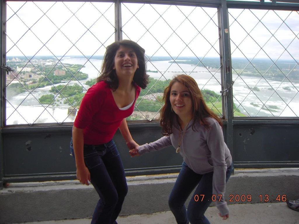 Two women holding hands and smiling on a high observation tower with a view of a river and cityscape in the background.