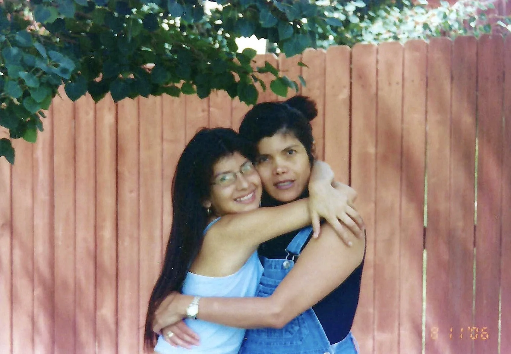 Two women hugging in front of a red wooden fence and green foliage, smiling and looking at the camera.