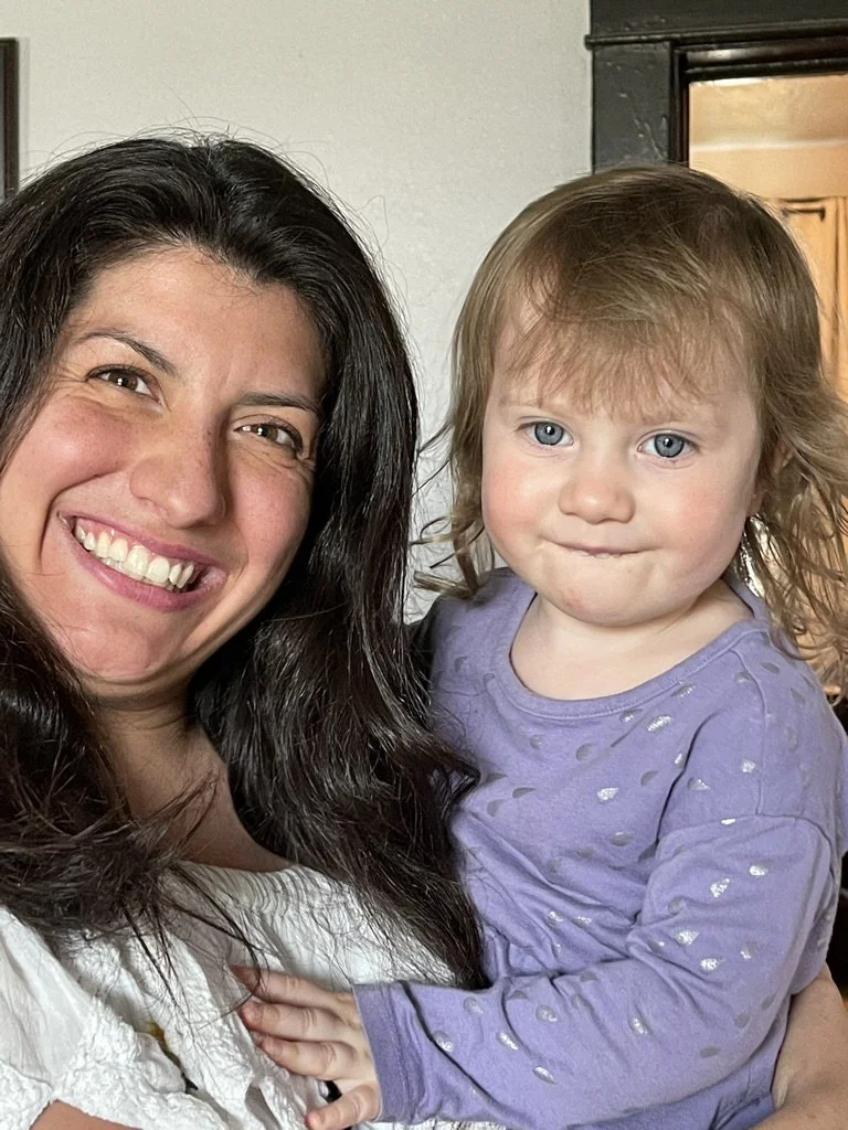A woman with long dark hair smiling and holding a young girl with light brown hair and blue eyes, wearing a purple shirt with small silver dots.