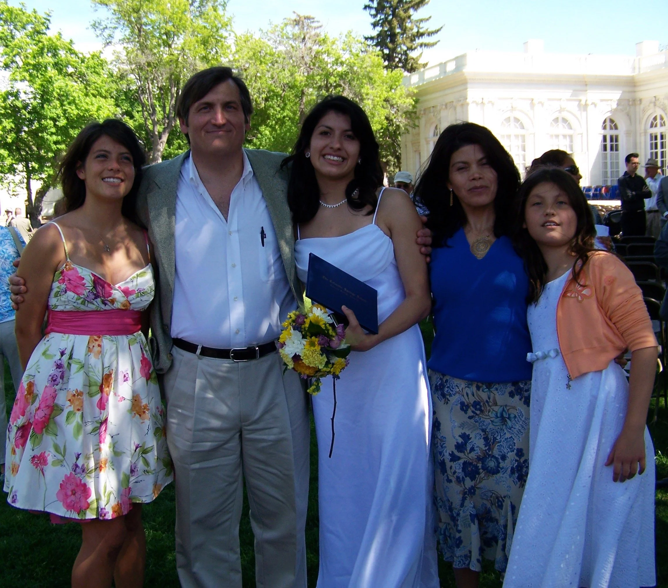 Group of five people standing outdoors on a sunny day, with a large white building and green trees in the background. The group includes three women, one man, and one child, all smiling and dressed in semi-formal attire. The woman in the center is ho