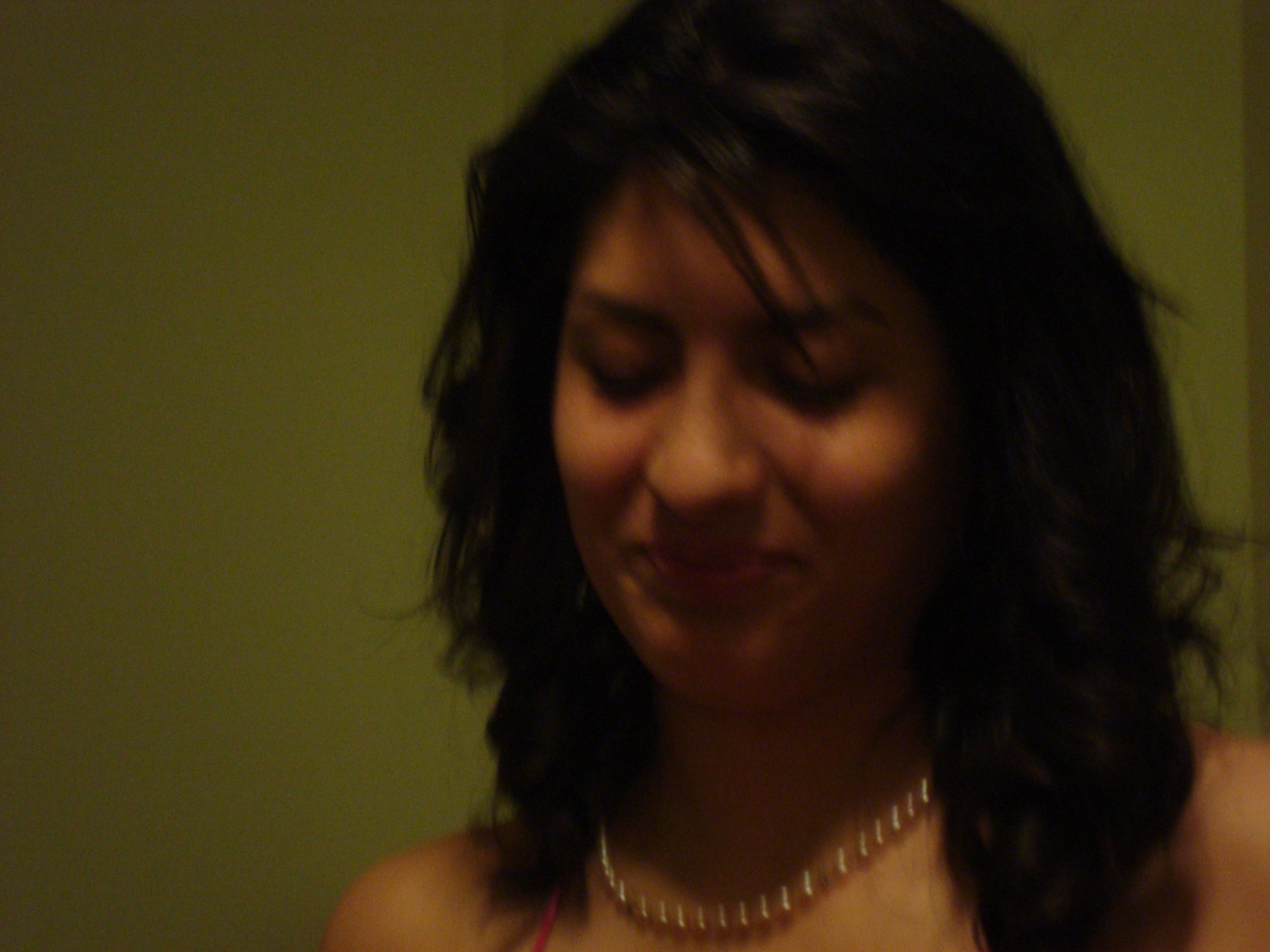 A woman with dark hair and a pearl necklace, smiling and looking down.