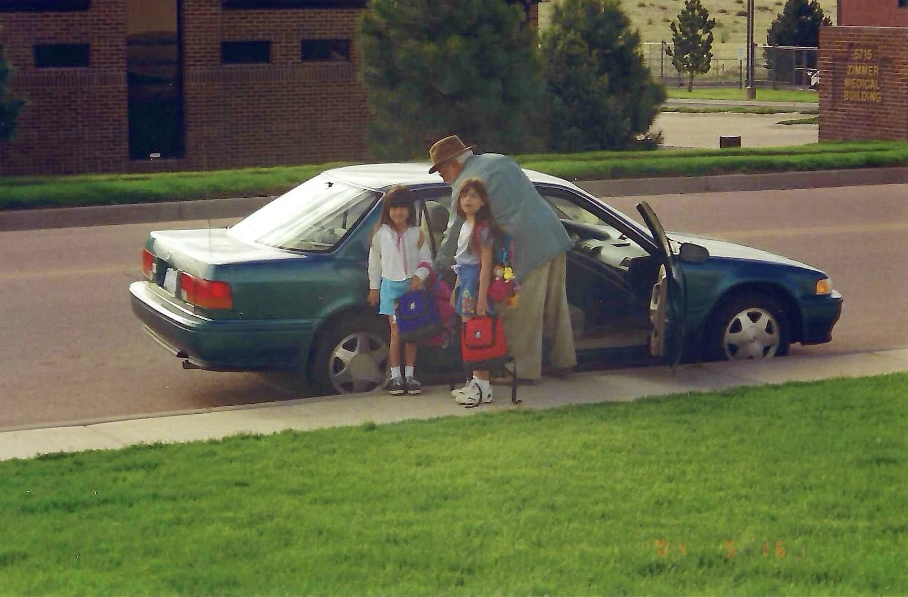 An elderly man helps two young girls with their backpacks next to a green car on a sidewalk in front of a brick building. The man is bending down, wearing a hat, gray jacket, and khaki pants, while the girls are holding their backpacks and the car do