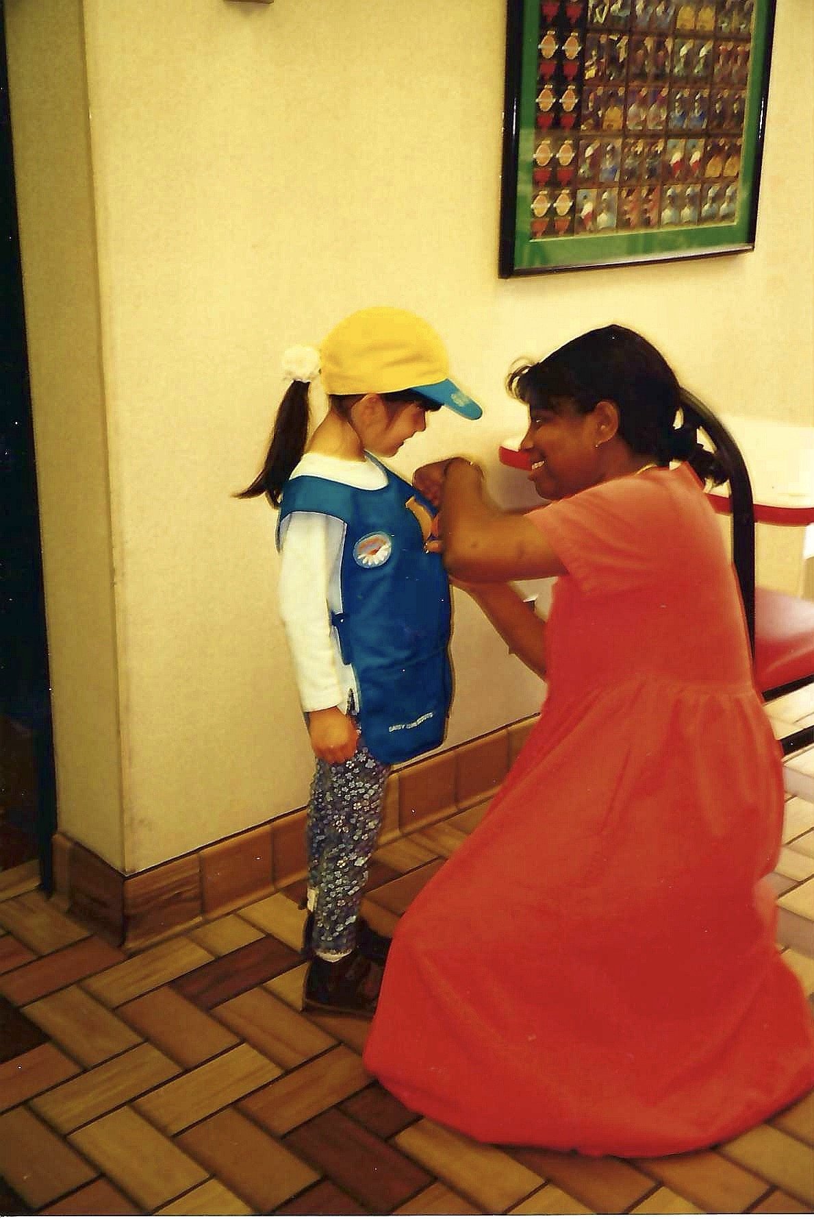 A woman in a red dress showing a young girl in a blue jacket and yellow hat how to do a handstand indoors, with a framed picture on the wall behind them.