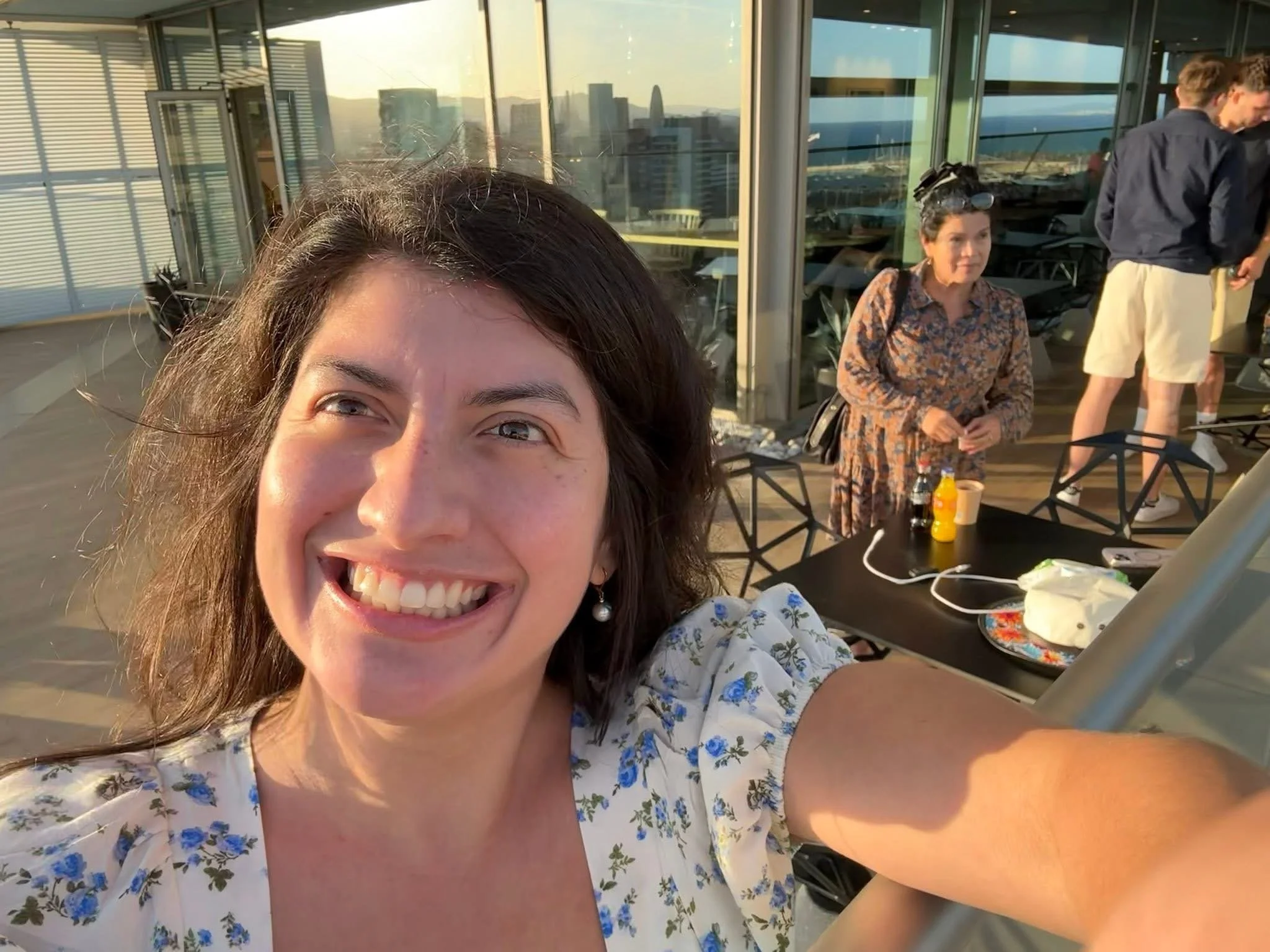 A woman smiling for a selfie on a balcony during sunset, with a city skyline in the background. There are several people in the background, including a woman standing near a table with drinks and snacks, and two men talking to each other.