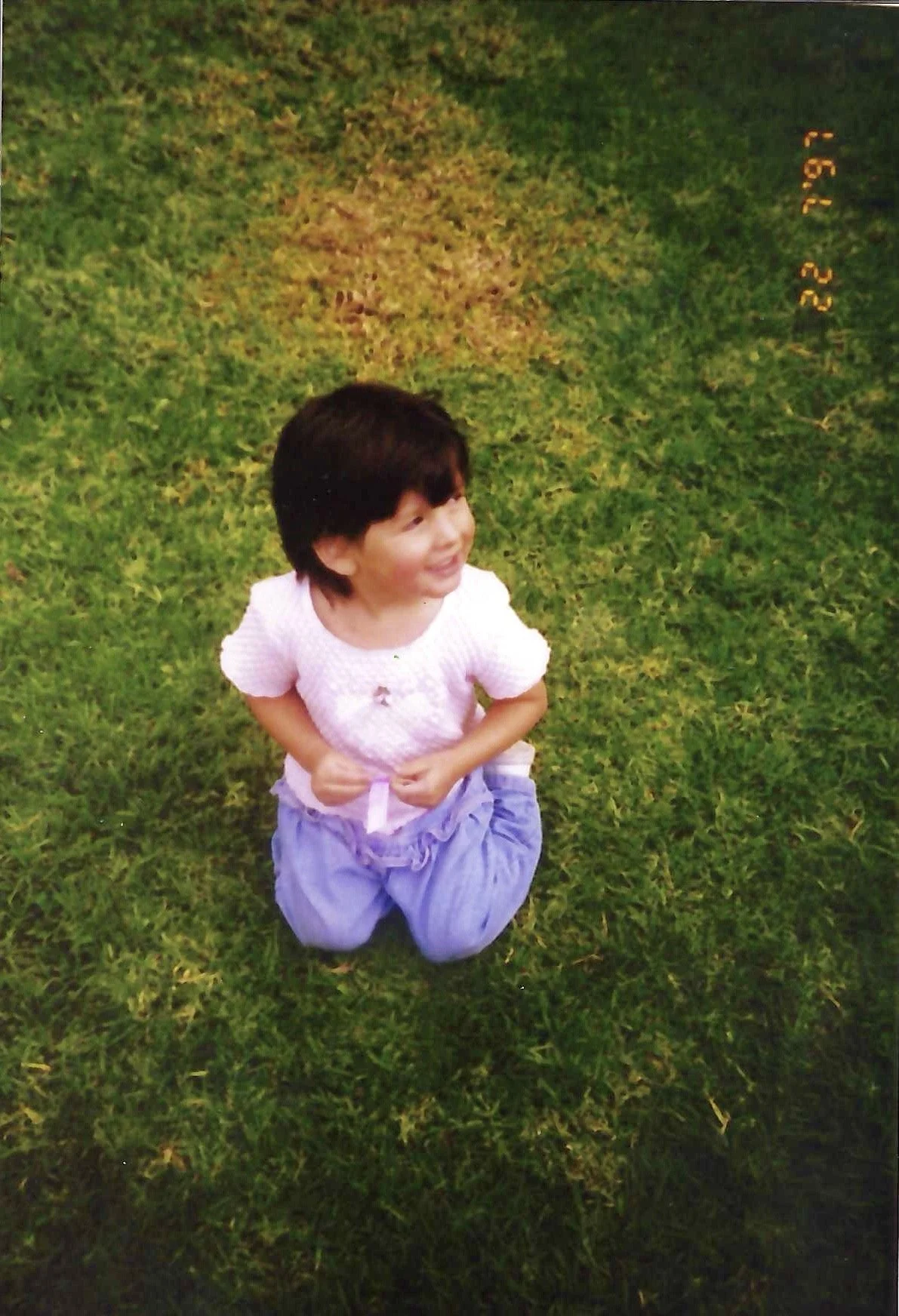 A young girl with short dark hair kneeling on grass, smiling and looking upward. She is wearing a light pink top and purple pants.