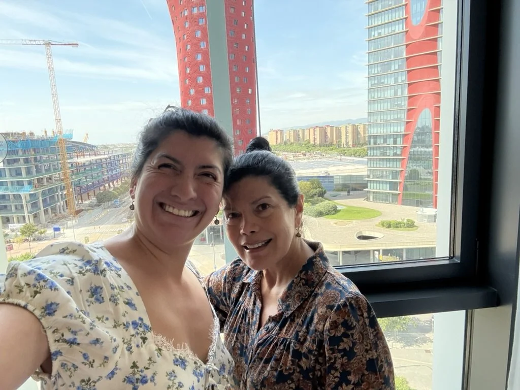 Two women smiling while taking a selfie in front of a large window with a cityscape view, including a distinctive red high-rise building and a construction crane.