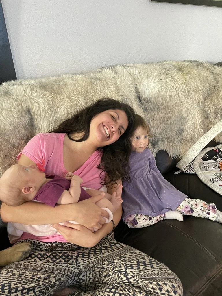 A woman sitting on a couch with two young children. She is smiling and holding a baby girl dressed in purple. Another girl in a purple dress and floral leggings is sitting beside her, looking to the side. The background includes a blanket, a pillow, 