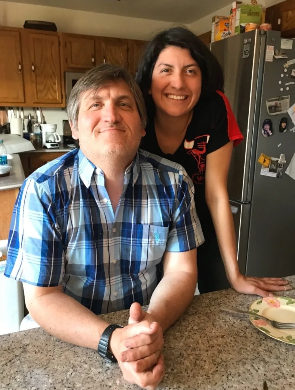 A man in a blue plaid shirt and a woman in a black shirt with red accents smiling in a kitchen, with the woman leaning on the counter behind him.