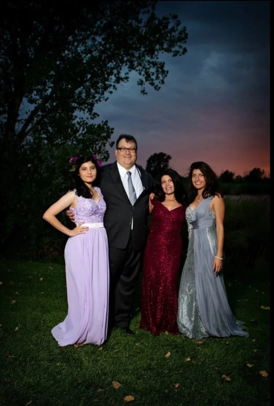 Four people dressed in formal attire standing outdoors during sunset, with trees and a darkening sky in the background.