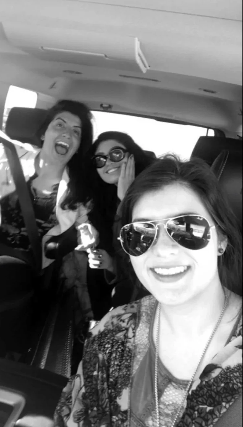 Four women sitting in a car, smiling and wearing sunglasses, with the woman in the foreground taking a selfie.