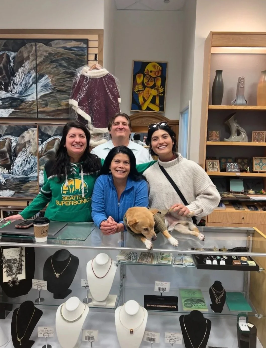 Four people and a dog inside a jewelry store with glass display cases, artwork, and shelves with decorative items.