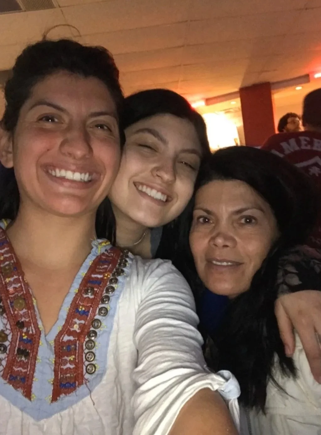 Three women are smiling and taking a selfie together in a dimly lit indoor setting, with warm lighting in the background.