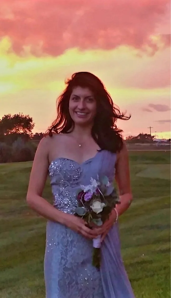 A woman in a purple dress holding a bouquet of flowers, smiling outdoors during sunset.