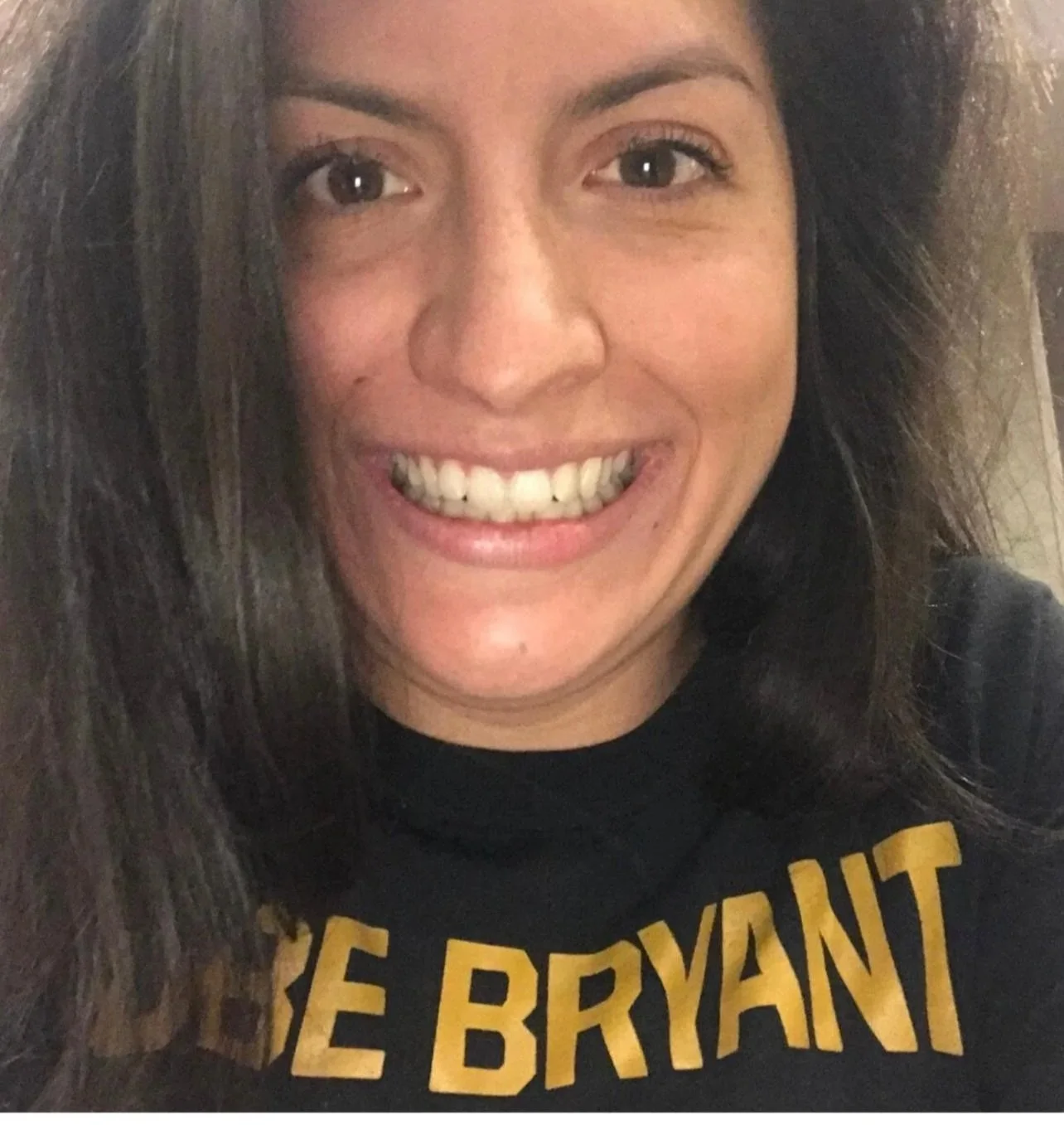 A smiling woman with long brown hair, wearing a black shirt with yellow text, in a close-up selfie.