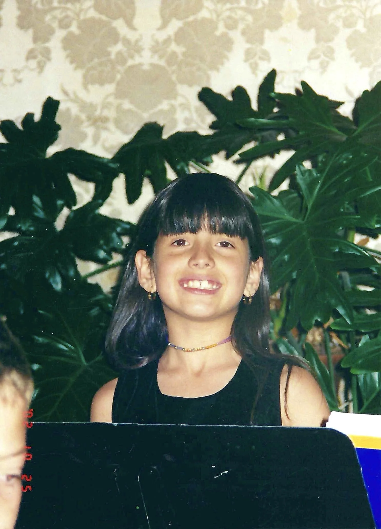 A smiling young girl with dark hair and bangs, wearing a black sleeveless top and colorful beaded necklaces, standing in front of green leafy plants and a patterned wall.