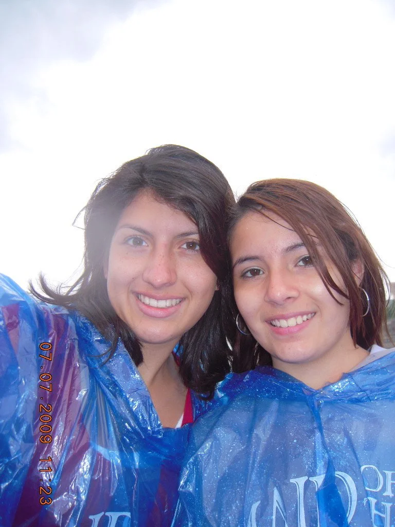Two young women smile for a selfie, wearing blue rain ponchos. One has dark hair, and the other has reddish-brown hair. The photo appears to have been taken during the daytime with an overcast sky in the background. The timestamp on the photo indicat