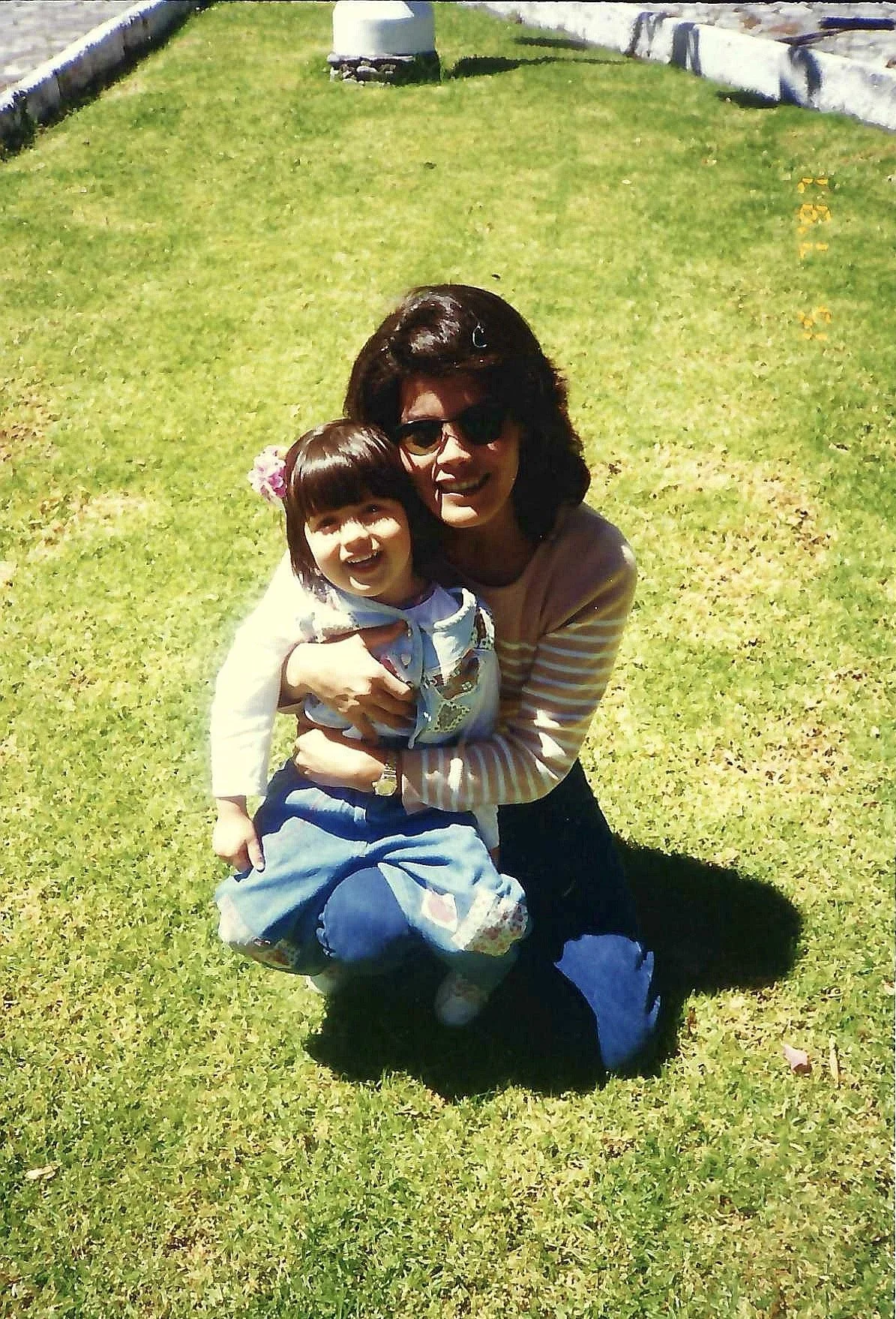 A woman with dark hair, sunglasses, and a striped beige and white long-sleeve shirt, hugging a young girl with a bob haircut, flower hair clip, and colorful clothing, both smiling on a sunny day on a grassy field.
