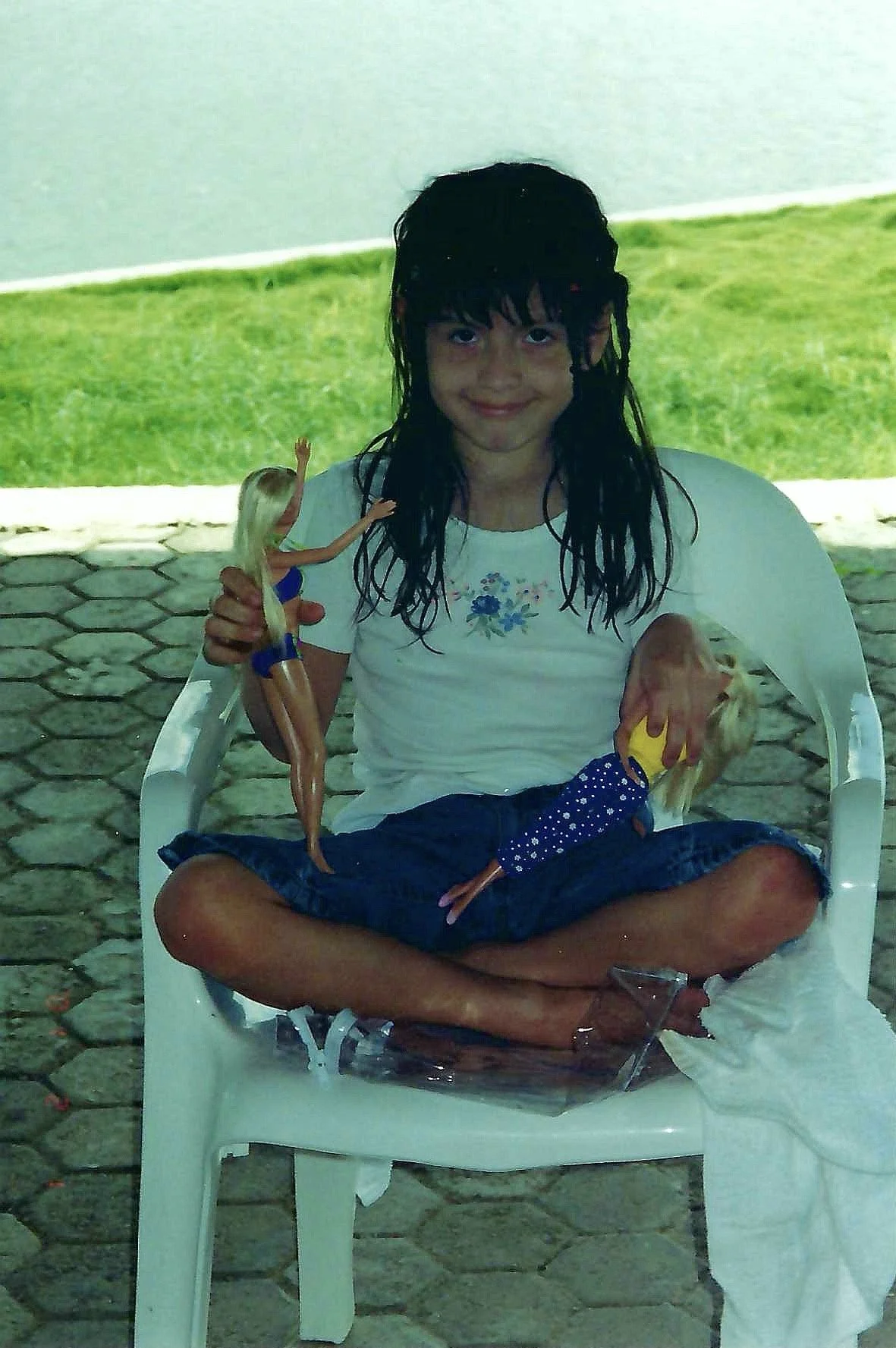 A girl sitting cross-legged on a white plastic chair outdoors, holding a large Barbie doll and a smaller doll. She has wet black hair, a white t-shirt with blue floral design, and a slight smile.