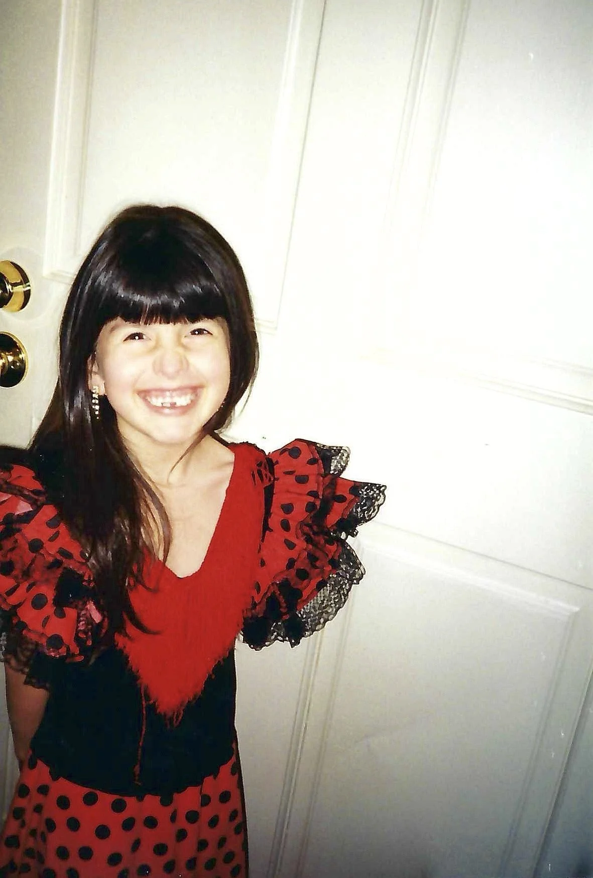 A woman smiling in front of a white door, wearing a red and black dress with polka dots and lace details.