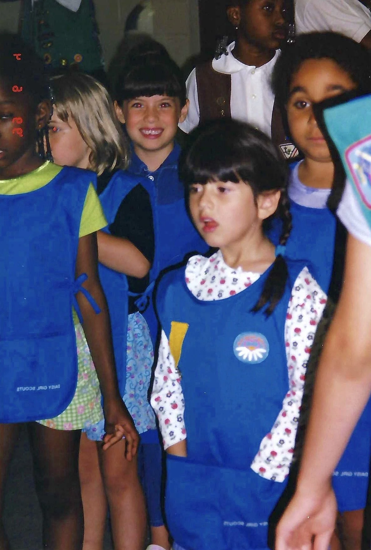 Group of children in school uniforms, holding hands, standing close together in a school or indoor setting.
