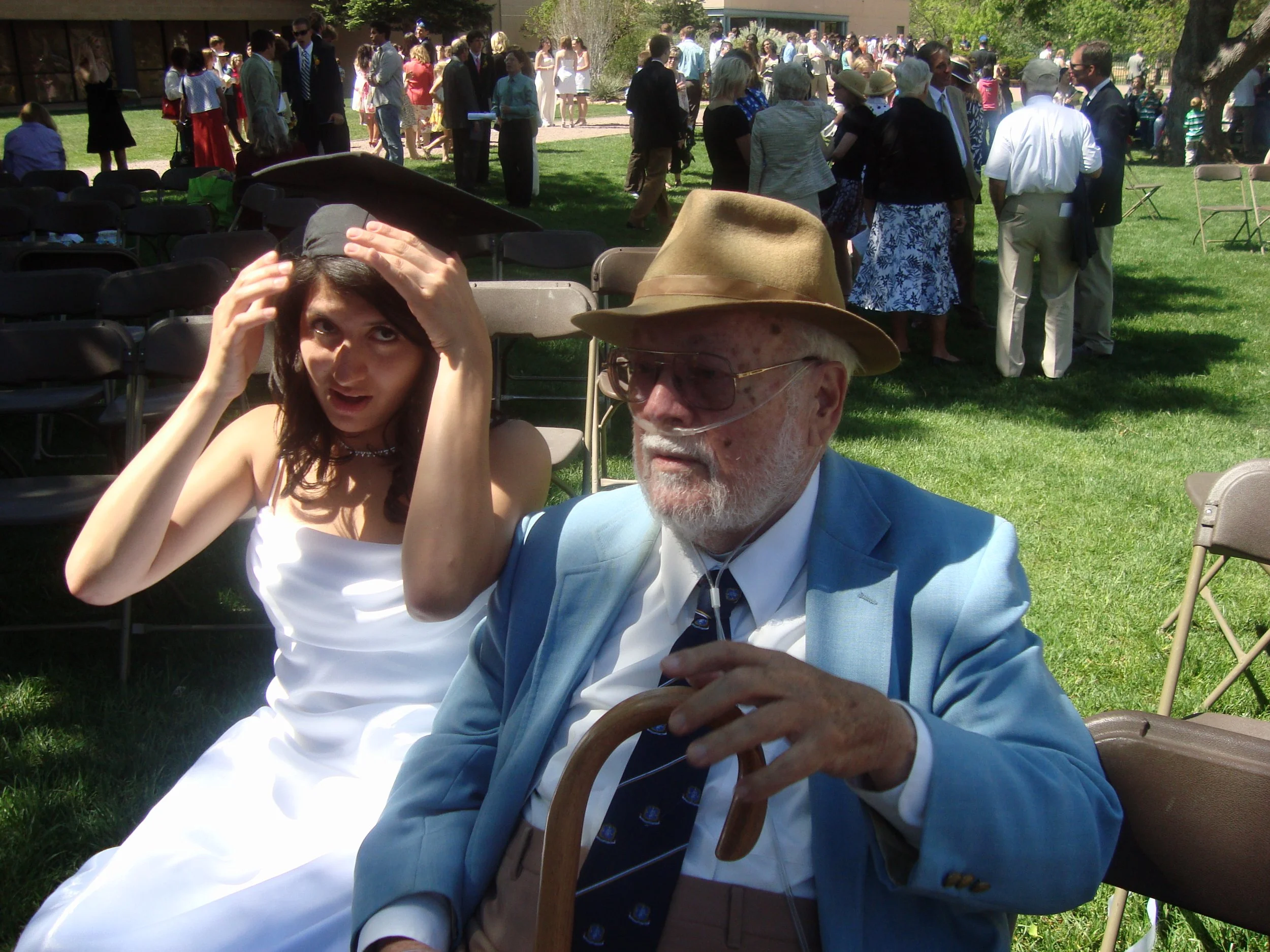 A young woman with dark hair and a middle-aged man with white hair and glasses wearing a tan hat are sitting outdoors at a large gathering in a park. The woman is adjust her hair with her hands, and the man is holding a wooden cane. Many people are s