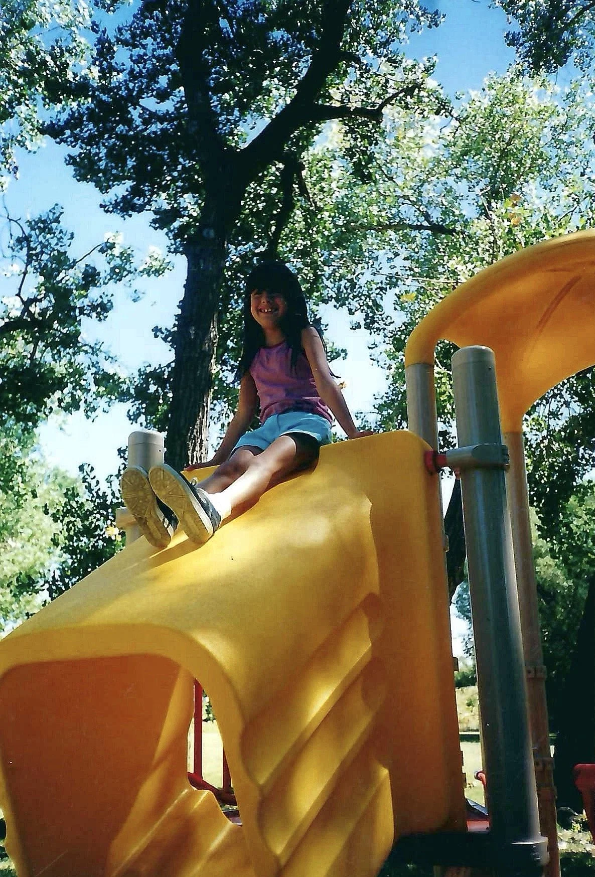 Young girl smiling and sitting at the top of a yellow playground slide in a park, surrounded by trees and clear blue sky.