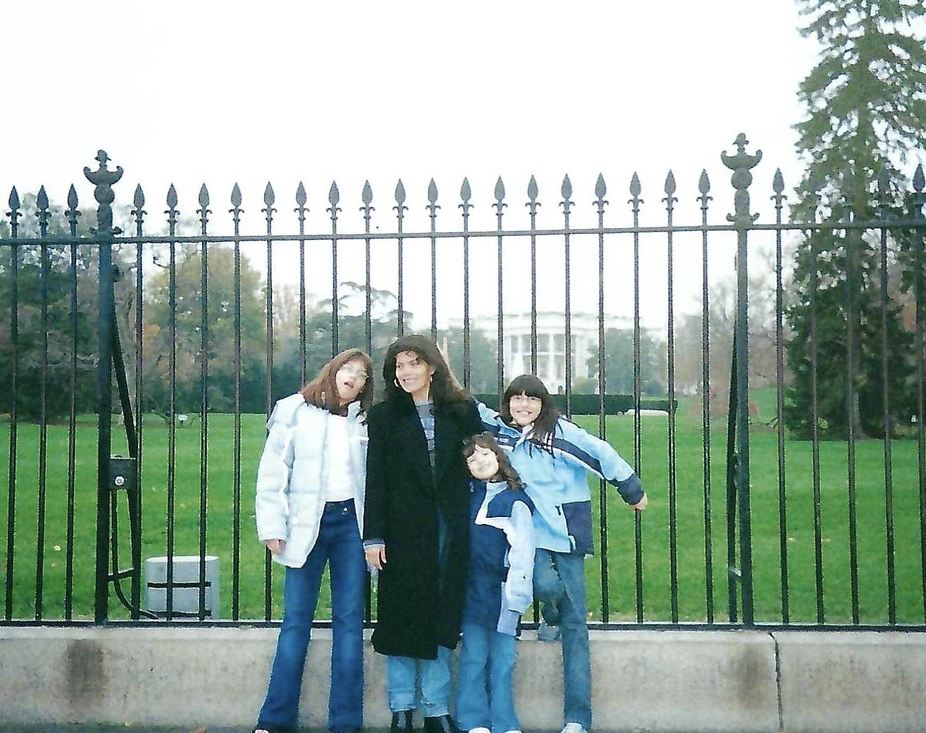 Four people, three women and one girl, standing in front of a black iron fence near the White House, smiling on a cloudy day.