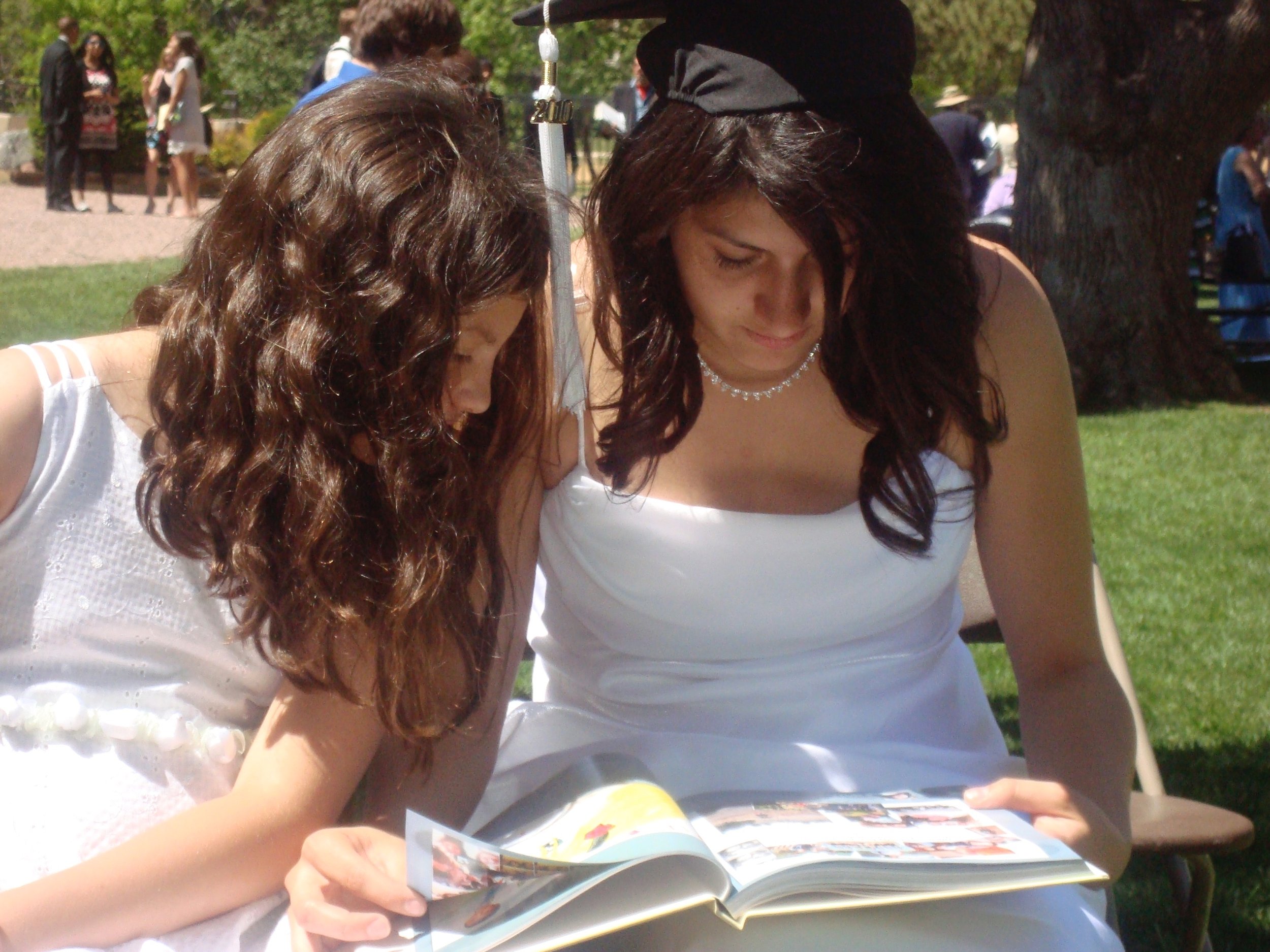 Two young women, one in a white dress and the other in a white top, are looking at a magazine together outdoors on a sunny day at what appears to be a graduation ceremony. The woman in the white dress is wearing a graduation cap.