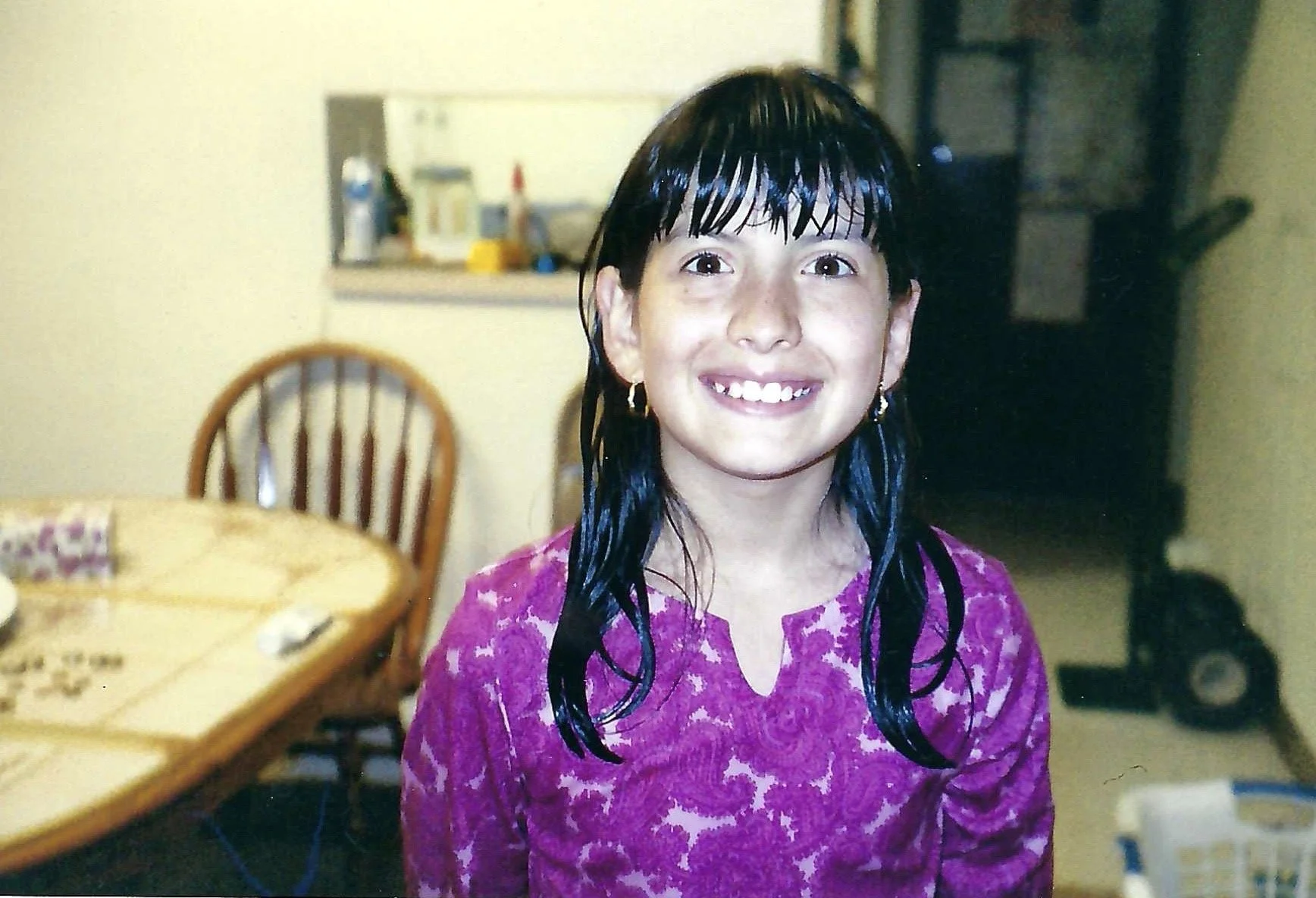 Young girl with wet black hair, smiling in a kitchen, wearing a purple patterned top.