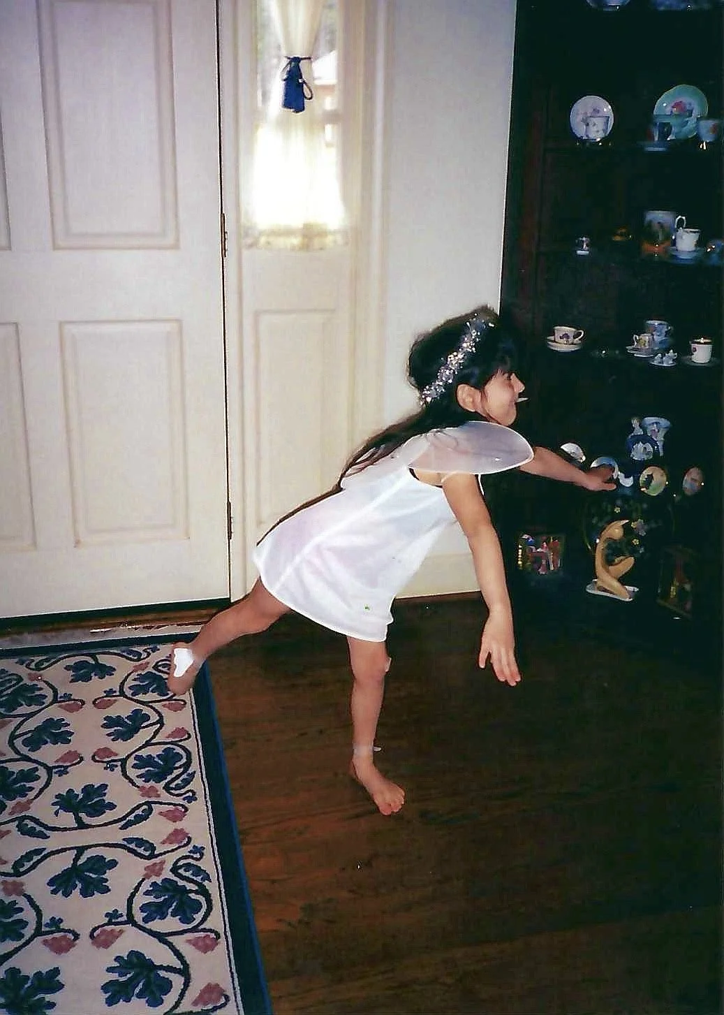 A young girl dressed as a fairy with a white dress, wings, and a silver headband, balancing on one foot with the other leg extended backward, reaching for a shelf of decorated plates and figurines in a home setting.