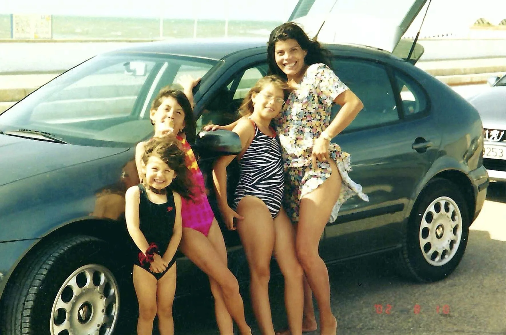 A group of five women and girls in swimsuits posing next to a black car with its hood open by the side of a road on a sunny day.