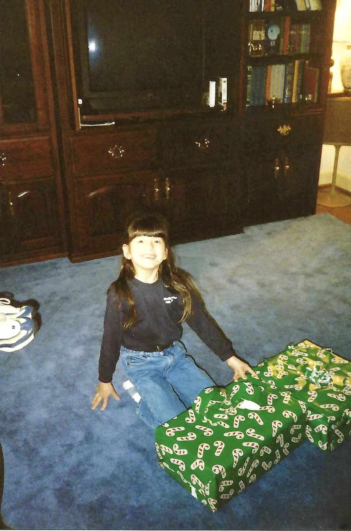 A girl sitting on a blue carpeted floor next to a large wrapped Christmas gift with a green wrapping paper and candy cane pattern. There is more wrapped presents and a pair of shoes nearby. In the background is a dark wooden TV stand with a televisio