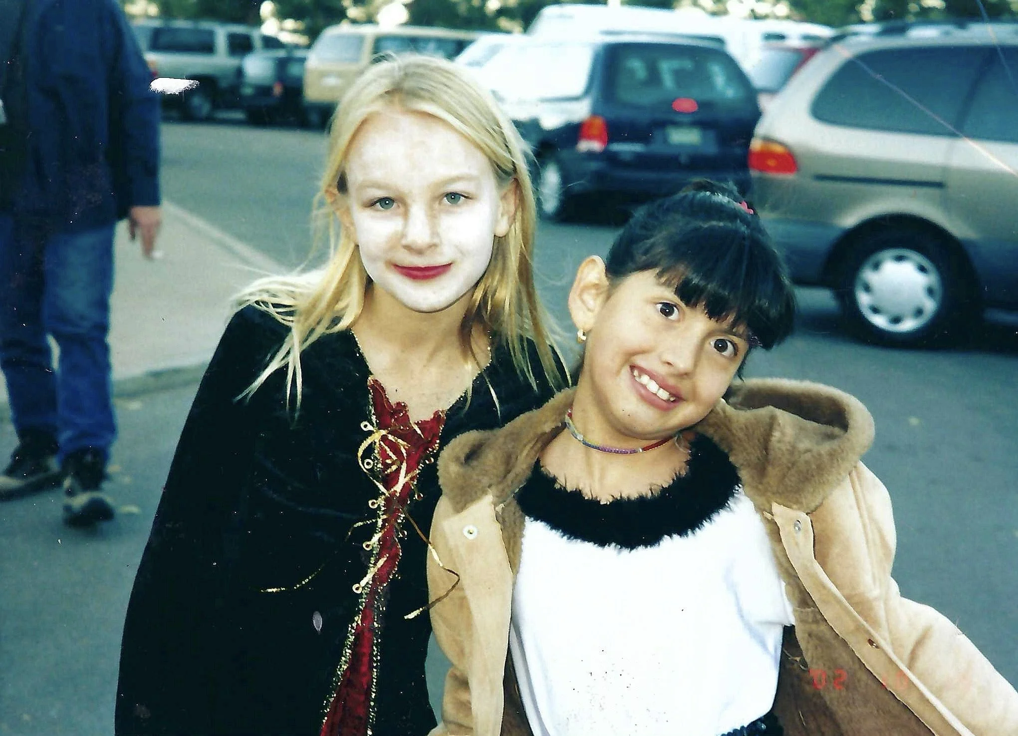 Two young girls smiling outdoors in a parking lot, one with blonde hair and the other with dark hair and bangs, dressed in casual winter clothing.