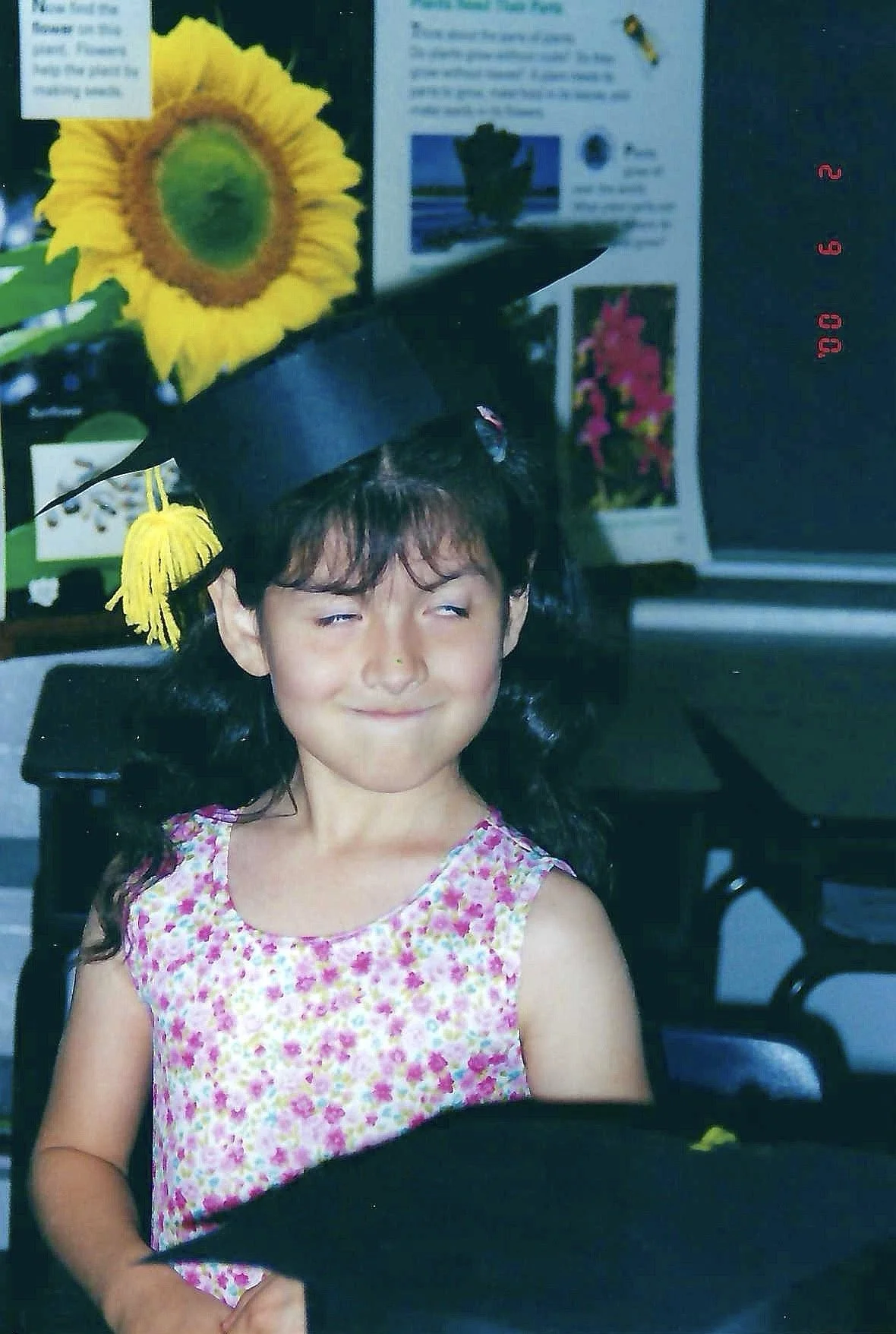A young girl with long black hair wearing a pink and white floral dress, smiling with her eyes half-closed, standing indoors in front of a bulletin board with pictures and text, wearing a black graduation cap decorated with a sunflower and butterflie