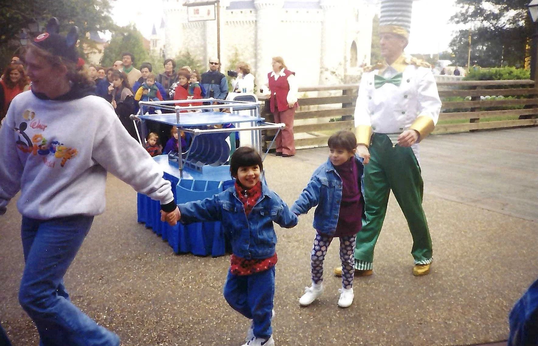 Children holding hands and walking in a circle outdoors at a theme park, with a man dressed in a colorful costume resembling a circus performer nearby.