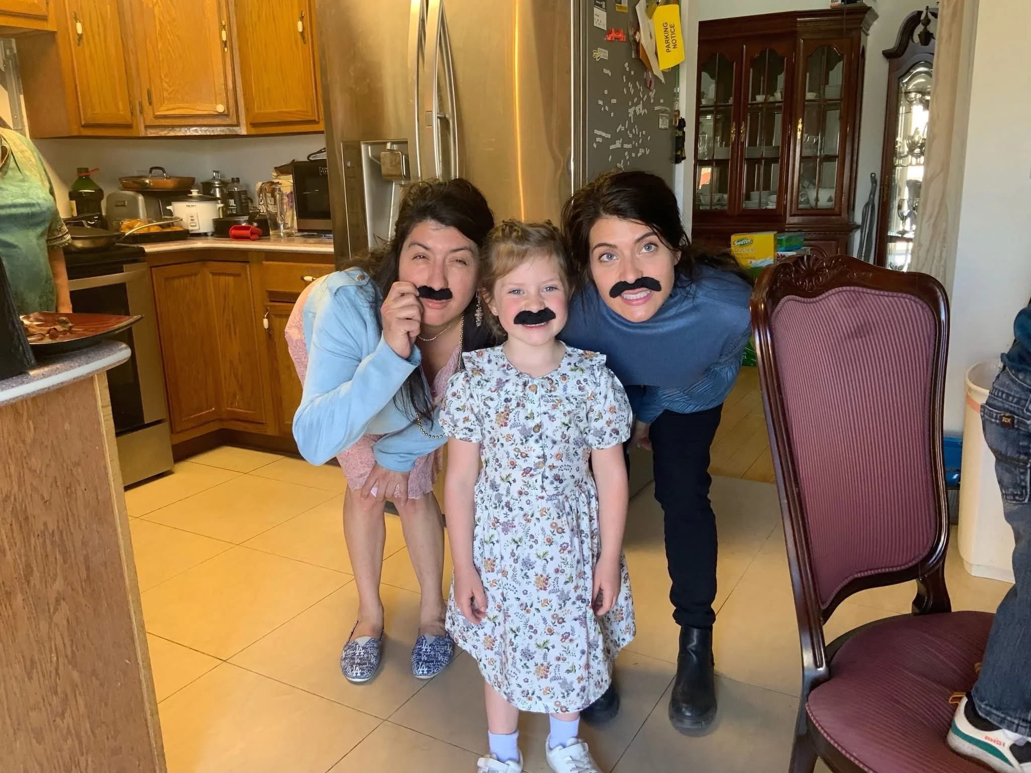 Three females, two adults and one young girl, with fake mustaches, in a kitchen. The girl is in the center, smiling, with long blonde hair, wearing a floral dress and sneakers. The woman on the left, with dark hair, is leaning in and smiling, holding