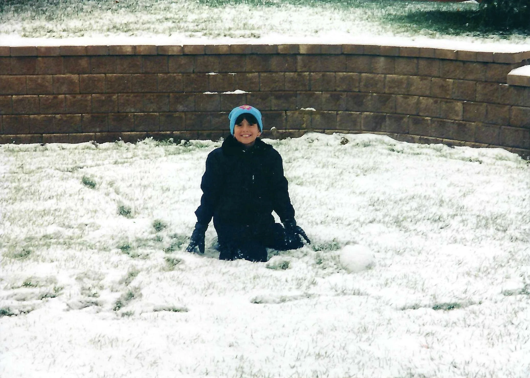 A smiling child wearing a black jacket and a blue winter hat with pink flowers, standing on snow-covered ground in front of a brick retaining wall with snow on top.