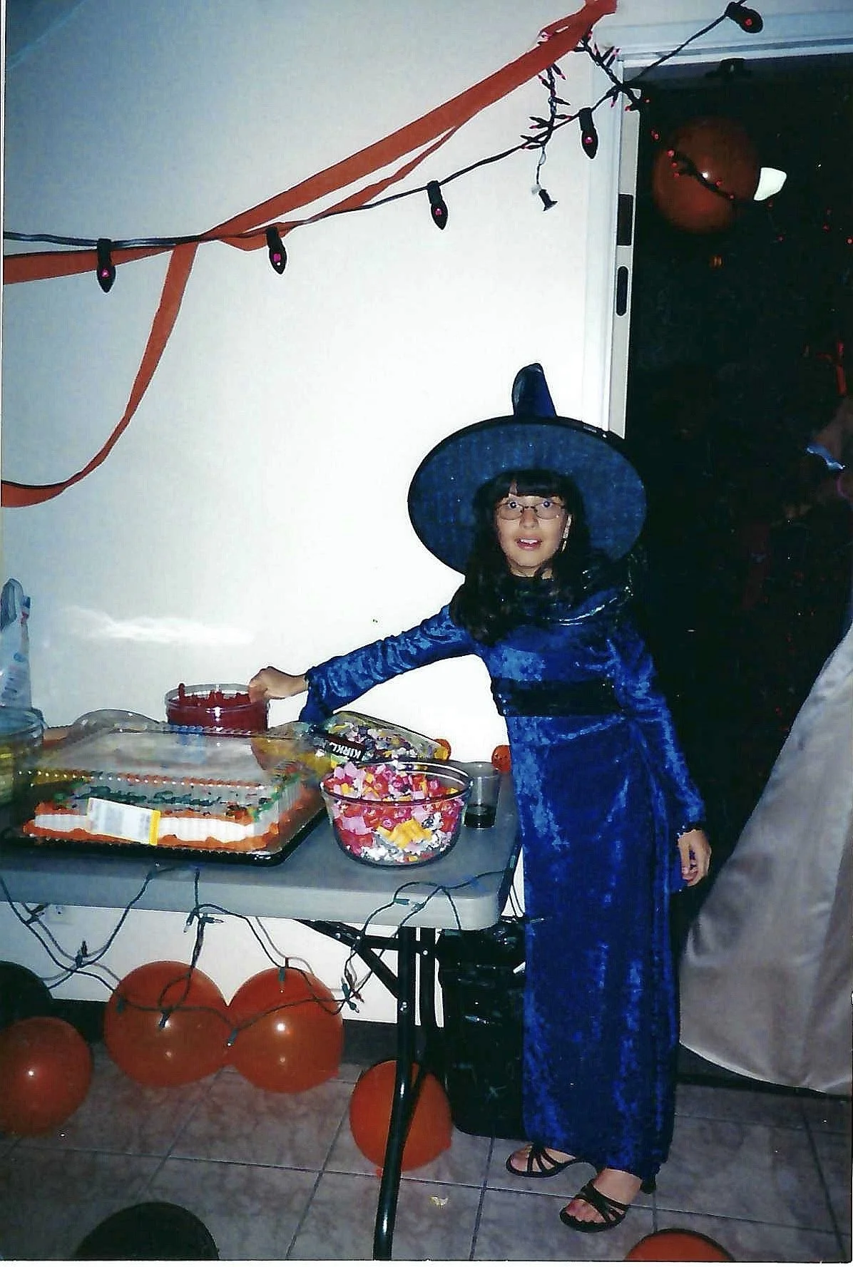 A girl dressed as a witch at a Halloween party, standing next to a table with candies and decorations. She is wearing a blue velvet witch costume and a large witch hat, with Halloween themed decor around her.
