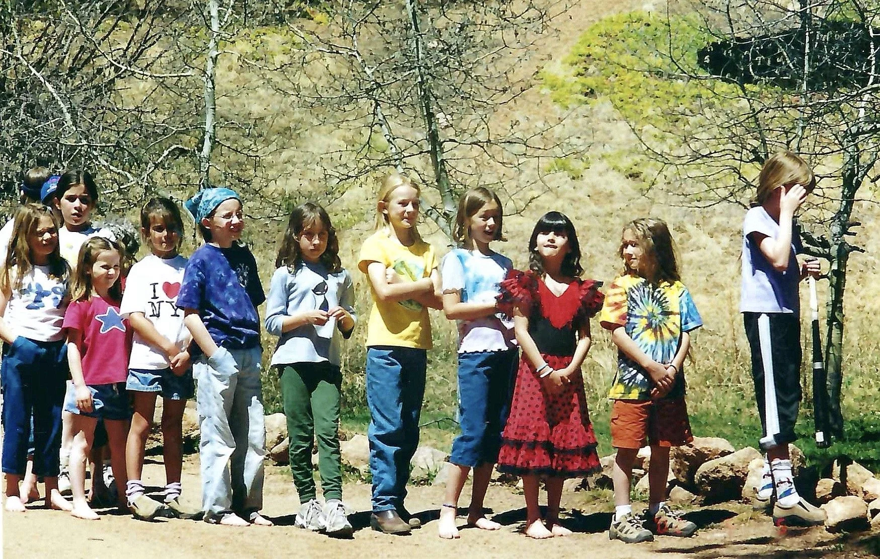 A group of children standing outdoors on a sunny day, some barefoot, with a wooded background, with a girl on the right adjusting her hair and holding a microphone.