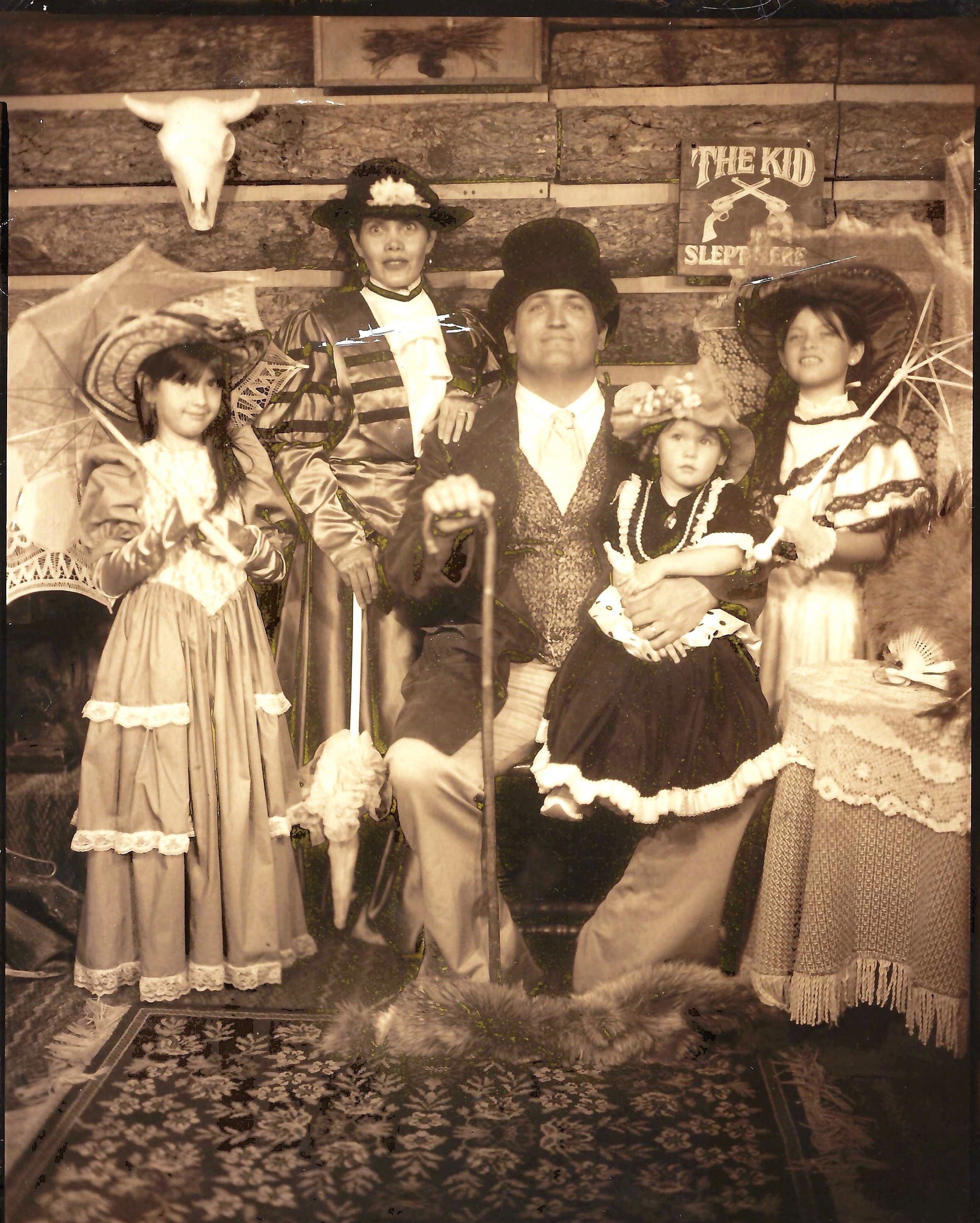 Vintage black and white photo of a family dressed in Old West costumes. Two women wear large, wide-brimmed hats and dresses with lace details, one holding a parasol. A man in the center wears a vest and tie, leaning on a cane. Three children are dres