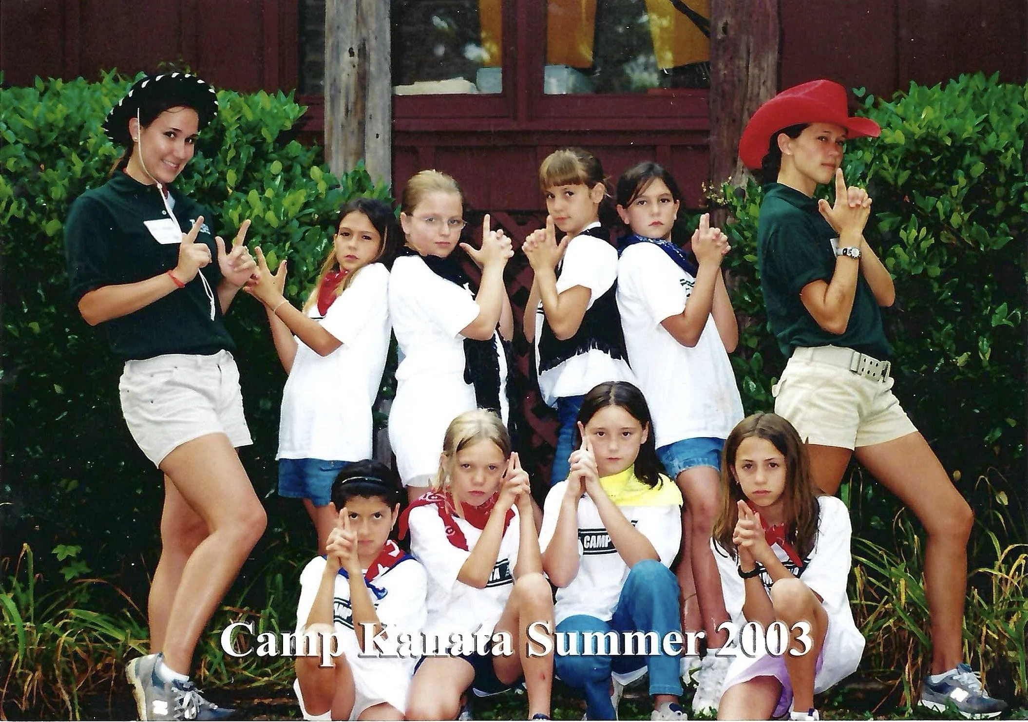 Group of children and women posing outdoors at a camp, with lush green bushes and a red wooden building in the background; some children are sitting, some women are standing with their thumbs up or making hand gestures; caption reads 'Camp Kauai Summ