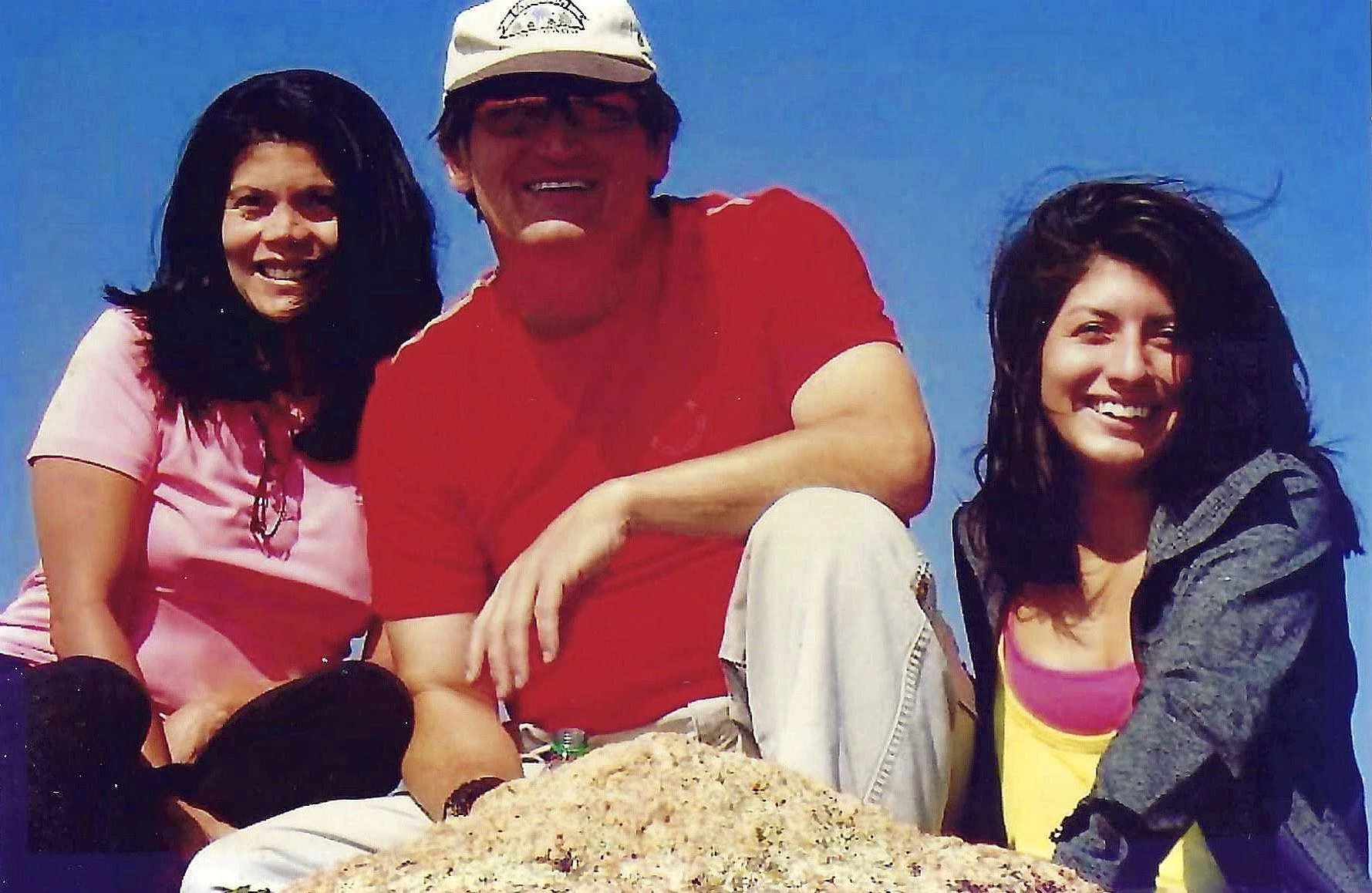 Three smiling young women and one younger man sitting outdoors against a clear blue sky. The man, in the center, is wearing a white cap, red shirt, and glasses, with his arm around the woman on his left. The woman on the left is wearing a pink shirt,