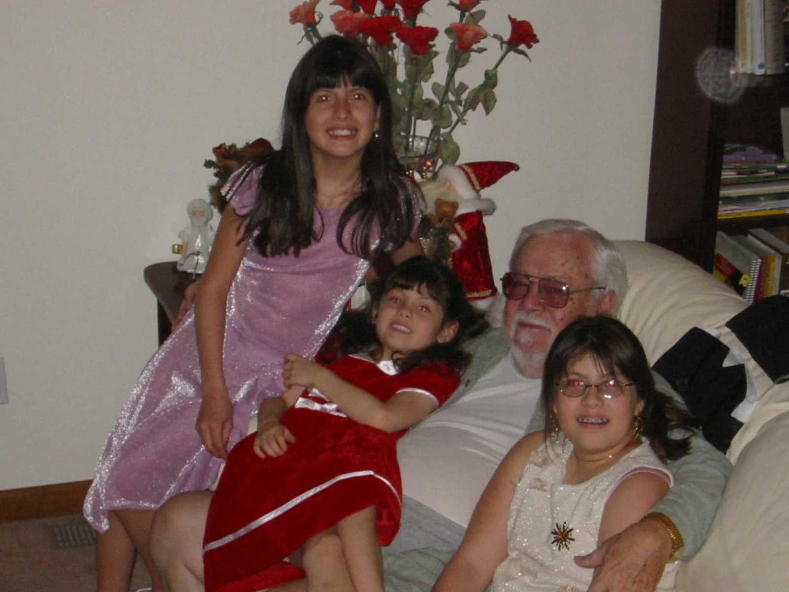 A family photo with four children and an elderly man, most likely a grandfather, in a living room with Christmas decorations including a small Christmas tree and a Santa figure. The children are dressed in festive attire, and everyone is smiling.