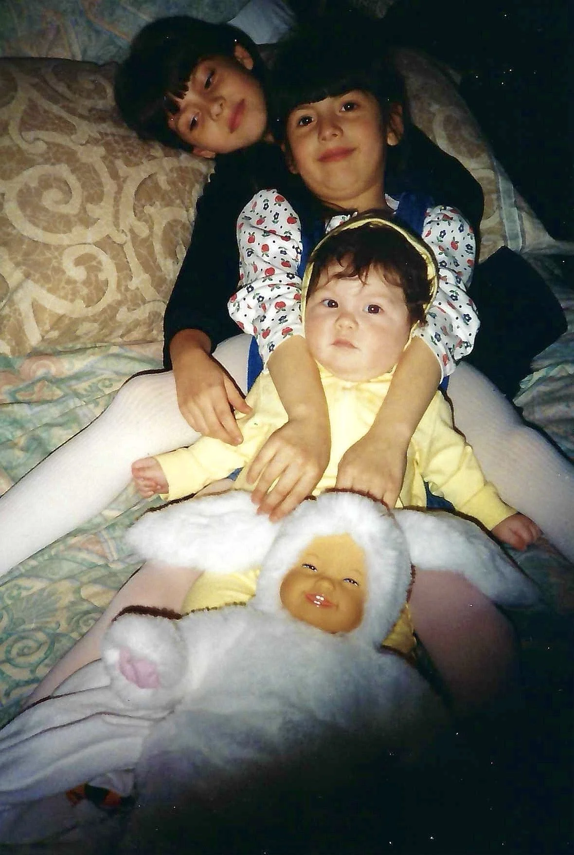 Three children lying on a patterned sofa, with a plush doll at the bottom of the image. The oldest girl has dark hair and is wearing a black and white outfit. The middle boy has short dark hair and is wearing a white shirt with colorful patterns. The