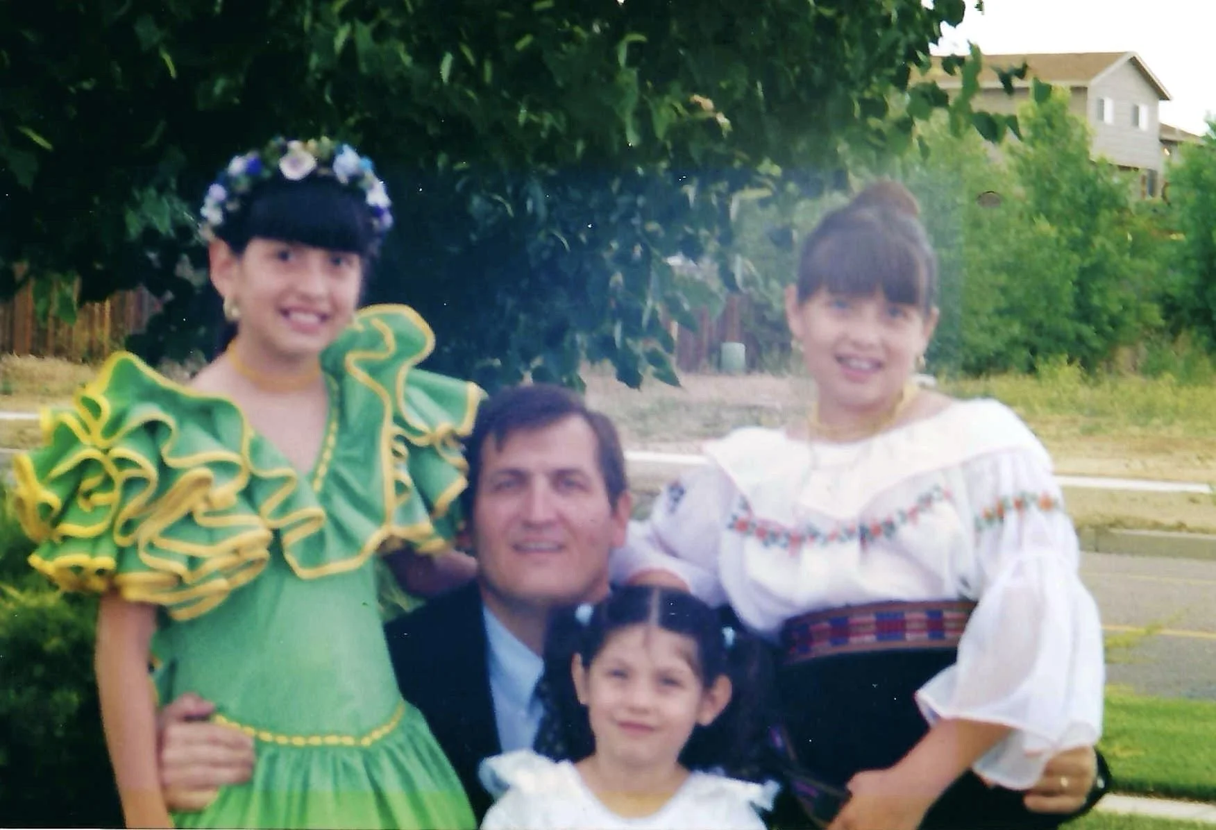 Group of people outdoors, some wearing traditional Mexican clothing, in front of green trees and houses.