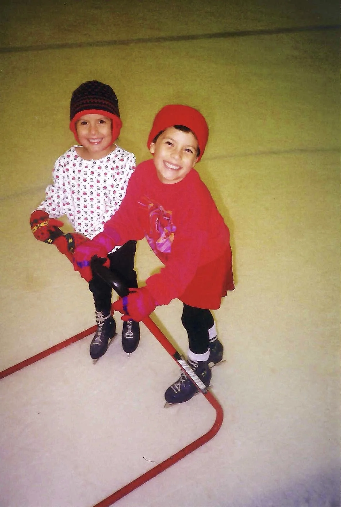 Two children ice skating indoors, holding onto a red skating aid. They are wearing winter hats, gloves, and warm clothing, and are smiling at the camera.