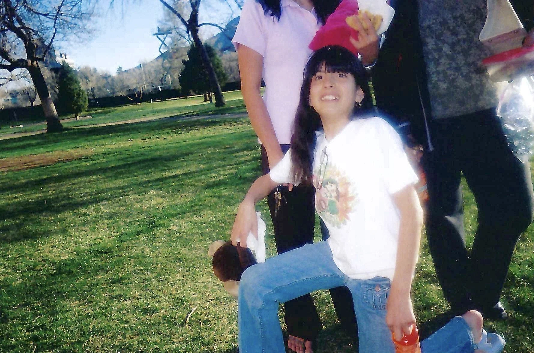 Two women and two children in a park, with trees and grass in the background. One woman is seated, and the other is standing near her. The children appear to be playing or interacting with each other.
