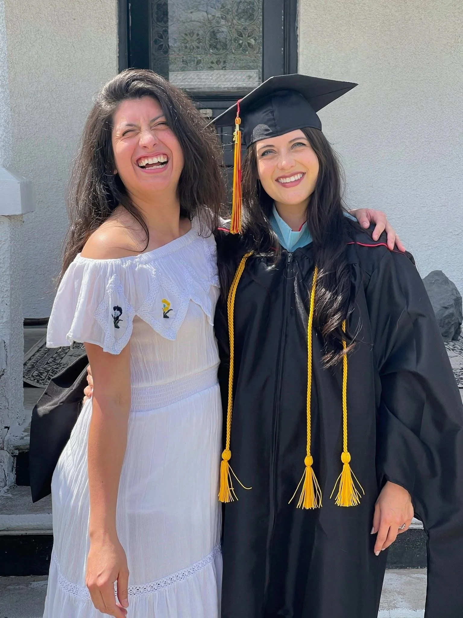 Two women, smiling joyfully, one wearing a white off-shoulder dress and the other in a black graduation gown and cap, standing outside in front of a light-colored wall with a window in the background.